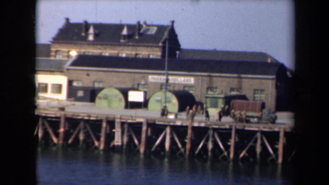 Wooden dock with industrial buildings, green tanks, a truck, and several people standing near the pier edge.