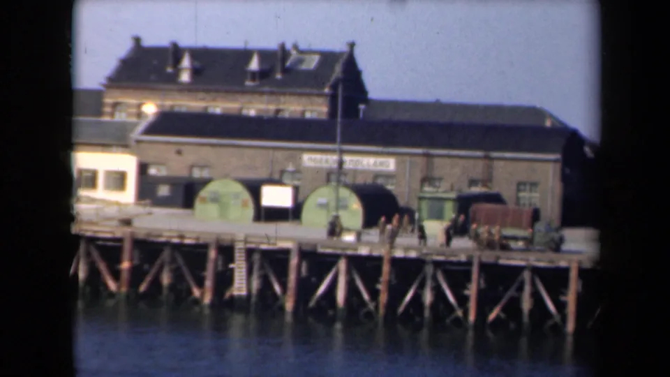 1946 Netherlands waterfront frame showing dock structures, green tanks, painted signage, a military-style truck, and people on the pier.