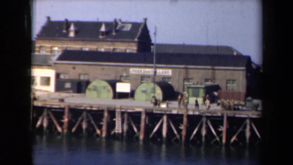 1946 Netherlands home-movie frame of a waterfront industrial building and pier, with people moving near green tanks and vehicles.