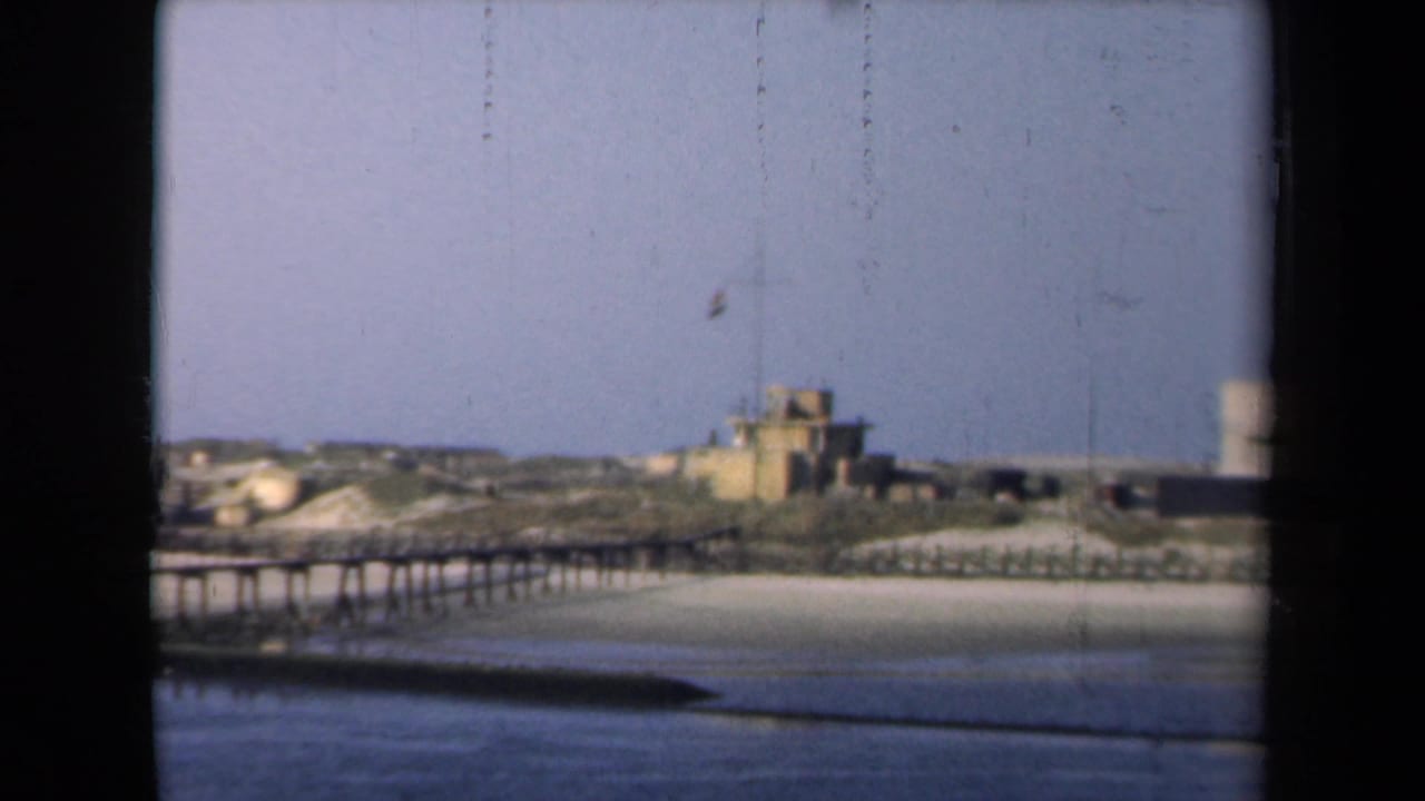 Wooden pier crosses calm water toward a sandy shoreline and small flag-topped building.