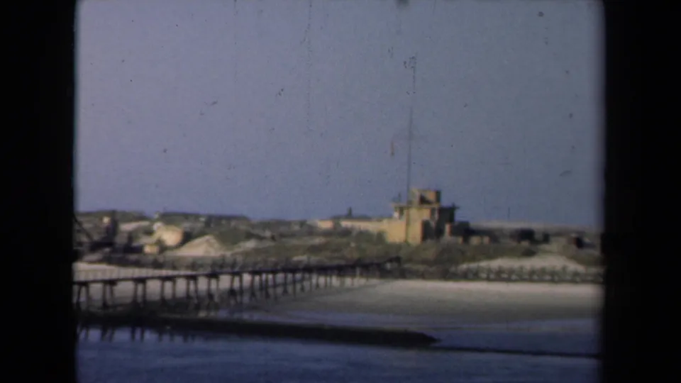 Soft 8mm view of a Netherlands harbor shoreline in 1946, showing a wooden pier, sandy bank, small flag-topped structure, calm water, and low distant buildings.