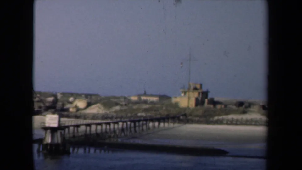 1946 Netherlands harbor shoreline with a wooden pier, small sign, sandy beach, calm water, low buildings, and a compact flag-topped structure in soft focus.