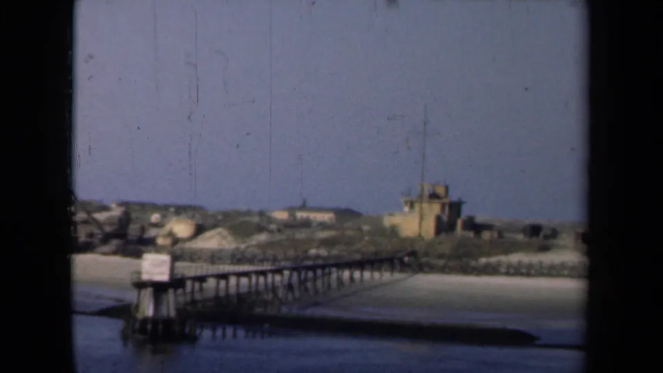1946 archival harbor view, supported by metadata, showing a wooden pier, calm water, sandy shore, low buildings, and clear sky.