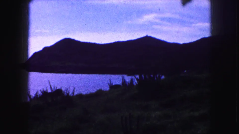 1959 8mm home-movie frame of a dark mountain ridge over blue water, with foreground grasses and window-like borders.