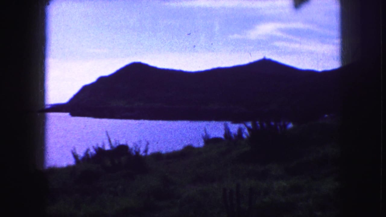 1959 8mm frame showing a dark coastal ridge, blue water, pale sky, foreground vegetation, and visible film-frame edges.