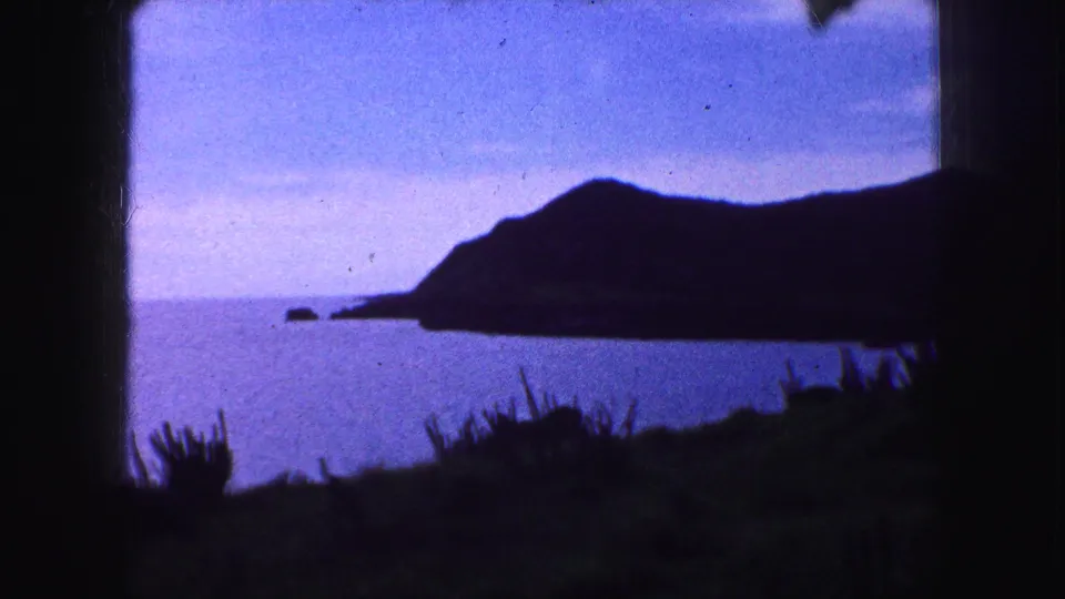 1959 8mm home-movie view of a dark headland and small offshore rocks above blue water, with foreground vegetation.