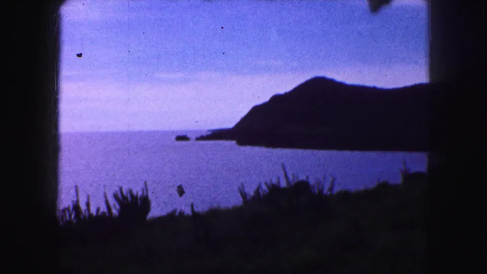 1959 8mm frame of a dark coastal mountain slope beside blue sea, with small rocks and foreground plants visible.