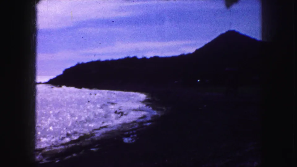 1959 8mm home-movie frame showing a shadowed beach, shimmering water, and a dark hillside or mountain form at the coast.