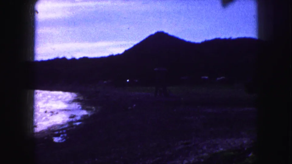 1959 8mm home-movie frame showing a dark beach below a silhouetted peak, with tiny distant people near the shoreline.