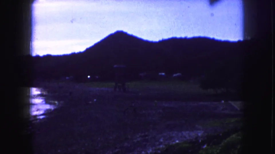 1959 8mm frame with a shadowed coastal foreground, distant people, reflective wet sand, and a dark ridge against the sky.