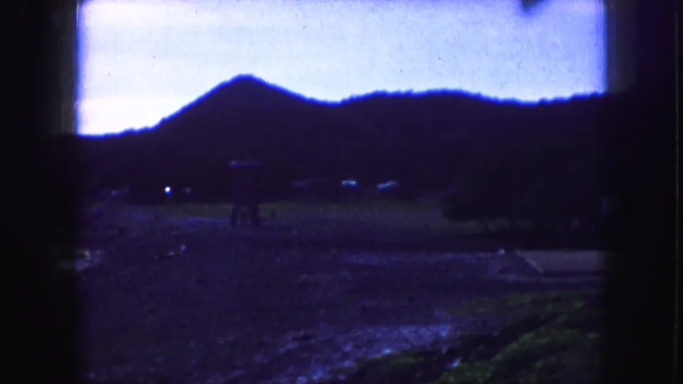 1959 Kansas City home-movie frame showing a dark wooded hillside, bright blue sky, wet foreground, and soft 8mm borders.