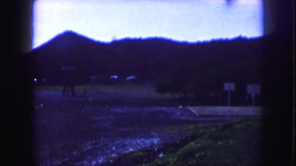 1959 Kansas City frame with a silhouetted hillside, wet creek-side ground, roadside signs, and distant vehicles in soft 8mm color.