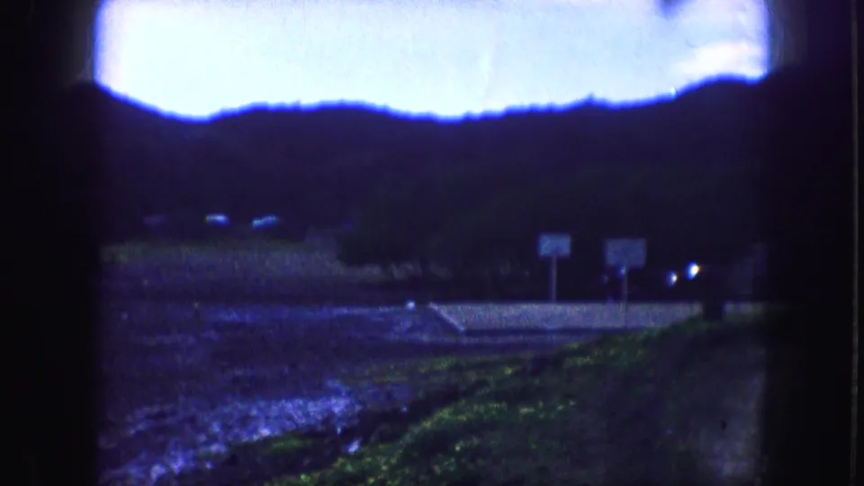 Blue-shifted 1959 Kansas City home-movie frame showing wet ground, a hillside, two roadside signs, and small vehicle lights.