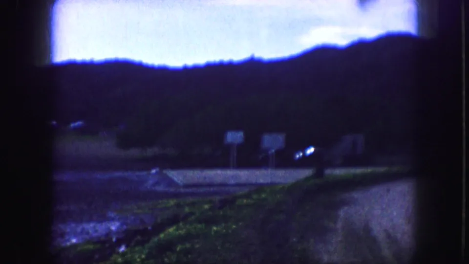 1959 Kansas City 8mm frame showing a dark hill, bright blue sky, two roadside signs, wet ground, and distant vehicle lights.