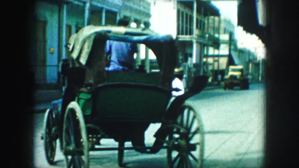 A green horse-drawn carriage with white wheels moves through a Kansas City street scene with early motor vehicles nearby.