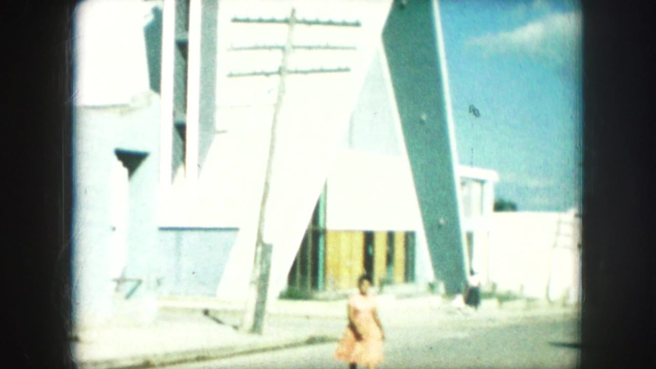 1959 Kansas City home-movie frame of a woman walking near a triangular modern building, with a utility pole and bright sky visible.