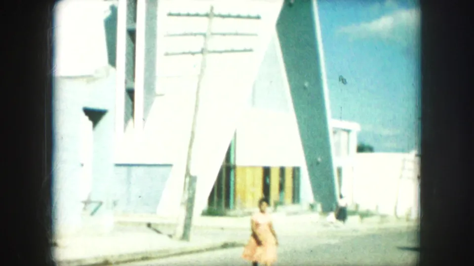 1959 Kansas City home-movie frame of a woman walking near a triangular modern building, with a utility pole and bright sky visible.