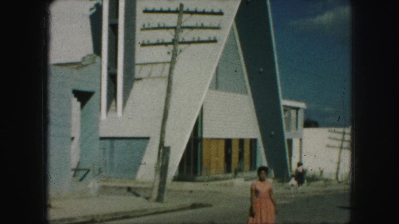 1959 Kansas City frame showing a woman in a pink dress walking toward camera outside a modern triangular building.