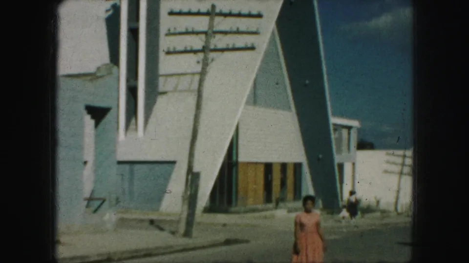 1959 Kansas City frame showing a woman in a pink dress walking toward camera outside a modern triangular building.
