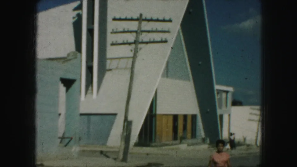 1959 Kansas City home-movie view with the woman low in frame and the modern building, pole, and sidewalk dominating the scene.