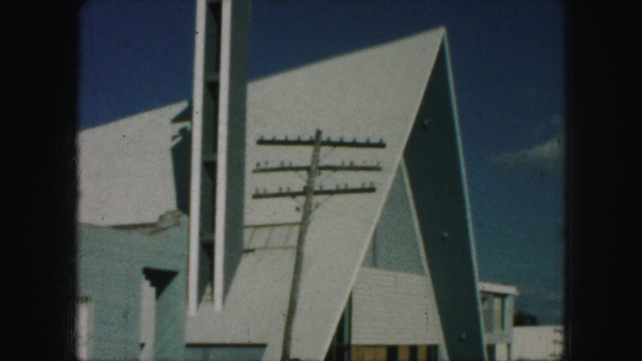 1959 Kansas City 8mm frame showing a white modern church exterior with angular roof planes, a tall cross-topped steeple, and a utility pole in front.