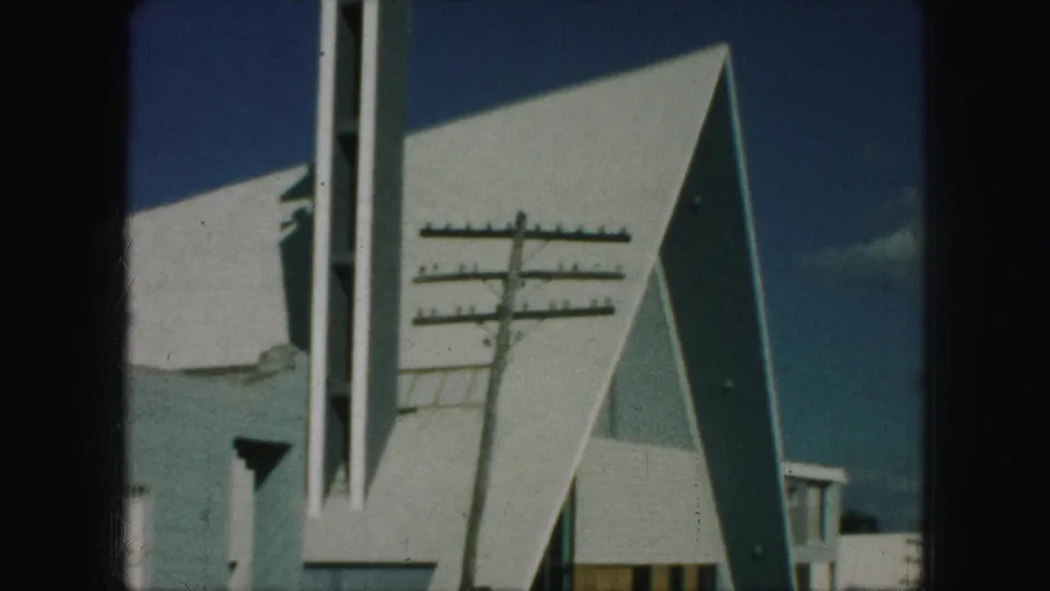 1959 Kansas City 8mm frame showing a white modern church exterior with angular roof planes, a tall cross-topped steeple, and a utility pole in front.