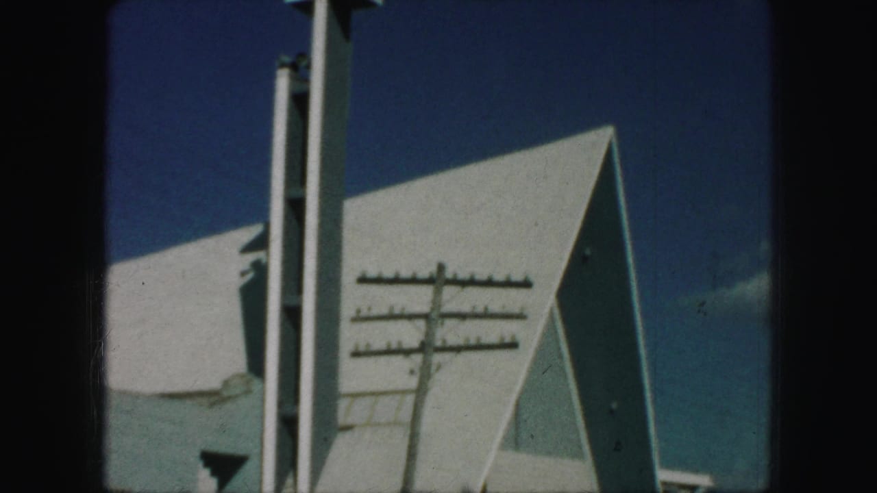 1959 Kansas City home-movie frame of a modern church roofline with a tall steeple and foreground utility pole against blue sky.