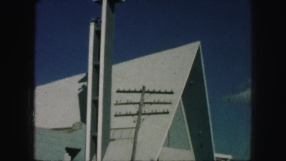 1959 Kansas City home-movie frame of a modern church roofline with a tall steeple and foreground utility pole against blue sky.