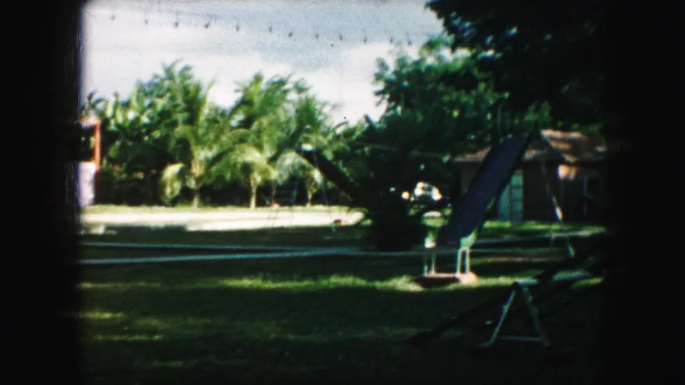 1959 Kansas City backyard playground scene with a metal slide, swing-set shapes, shaded lawn, trees, and residential buildings.