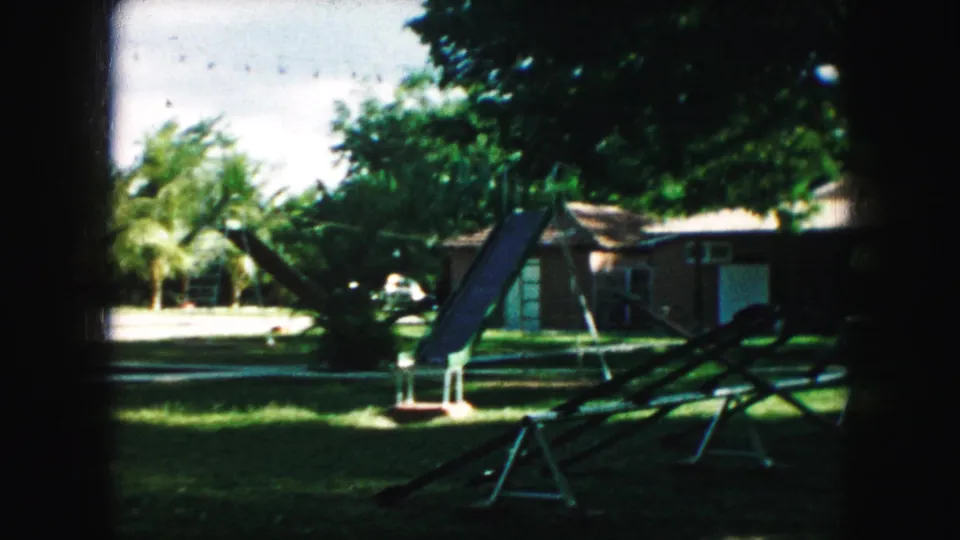 1959 Kansas City home-movie frame showing backyard play equipment, shaded grass, trees, and single-story residential buildings.