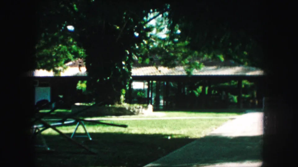 1959 Kansas City home-movie frame showing a shaded suburban lawn, house facade, porch area, and partial play structure.