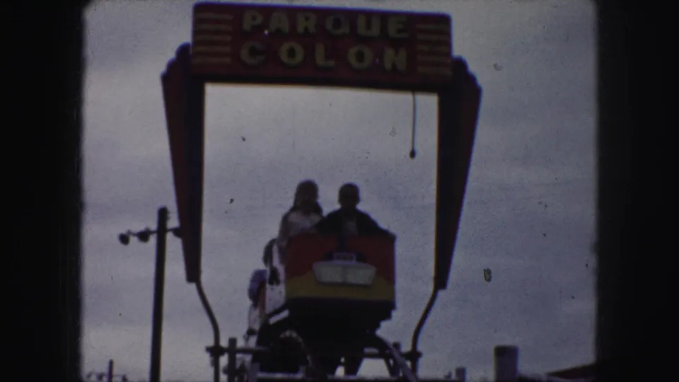 A coaster car carrying several riders moves toward the camera below the Parque Colon sign in 1959 Kansas City footage.