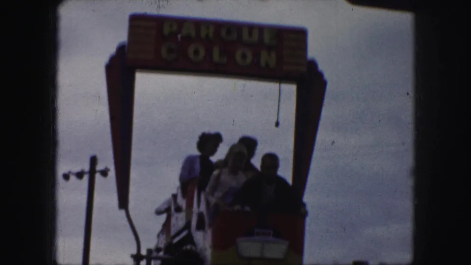 Several riders, including a woman near the front, pass under the Parque Colon sign on the roller coaster in Kansas City footage.