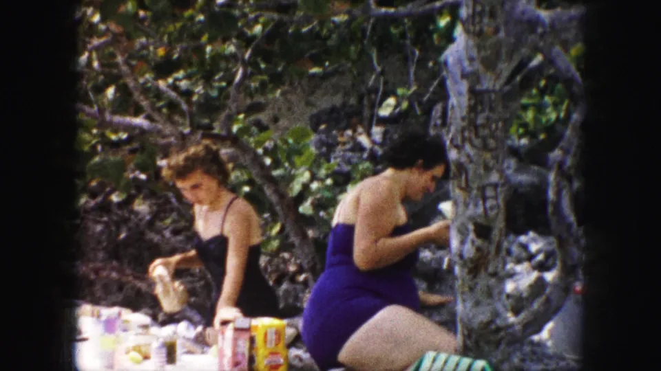 1959 Kansas City home-movie frame showing two women in one-piece swimsuits preparing food at an outdoor table in a shaded rocky setting.