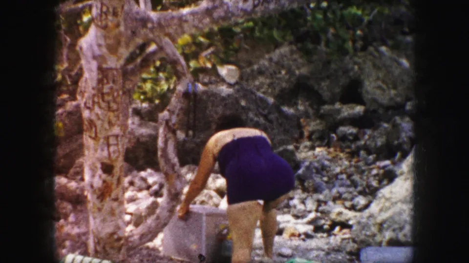 1959 Kansas City home-movie frame with a woman in a purple one-piece swimsuit reaching toward a container in a rocky shaded area.