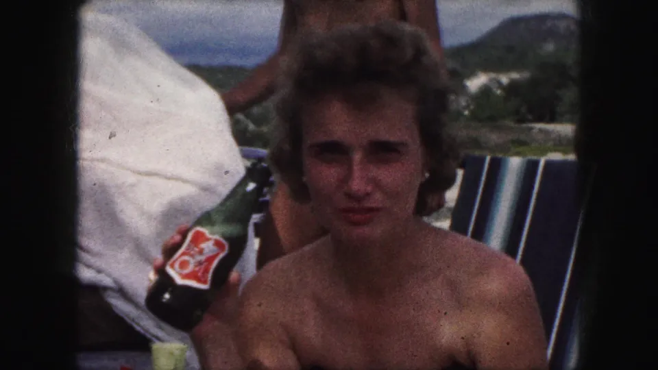 In 1959 Kansas City-area home-movie footage, a seated woman in a black swimsuit faces the camera while holding a labeled green bottle.