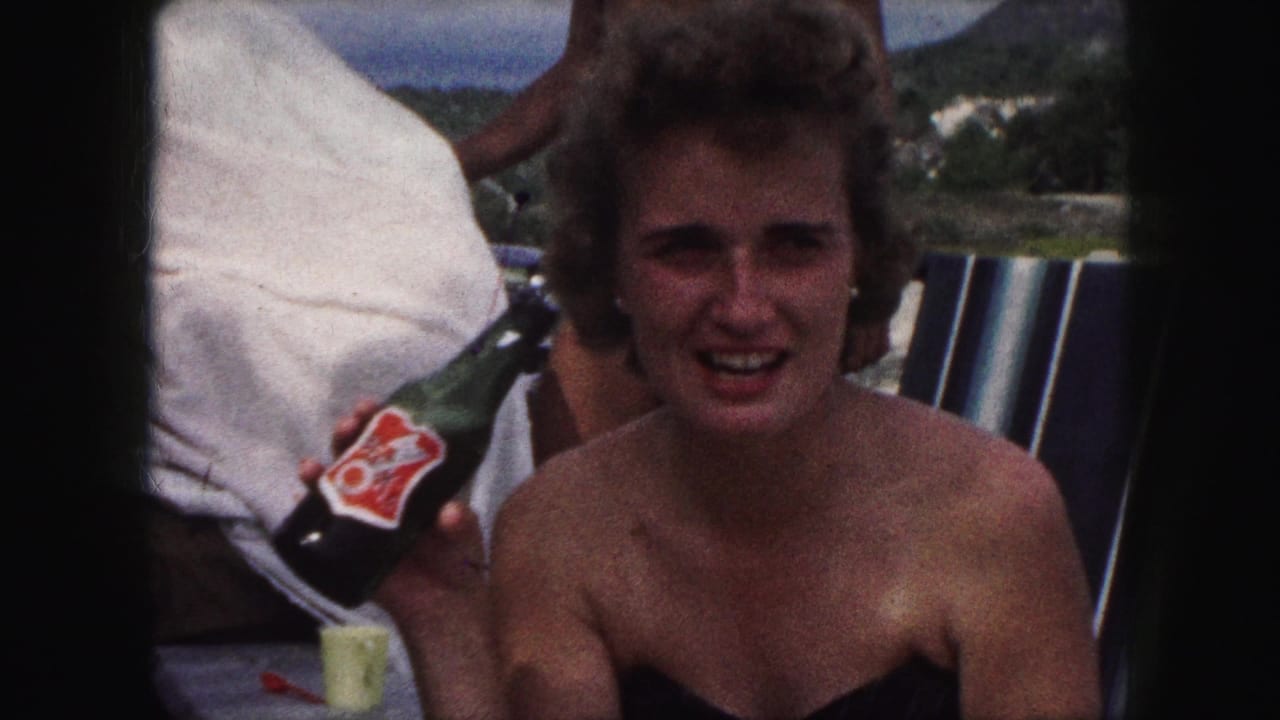 The woman smiles in the 1959 outdoor home-movie scene, seated near a striped chair with a labeled green bottle in hand.