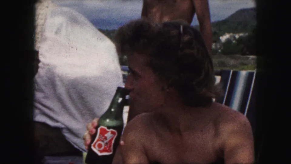 The 1959 Kansas City home-movie frame shows the woman drinking from a labeled green bottle beside a striped chair outdoors.