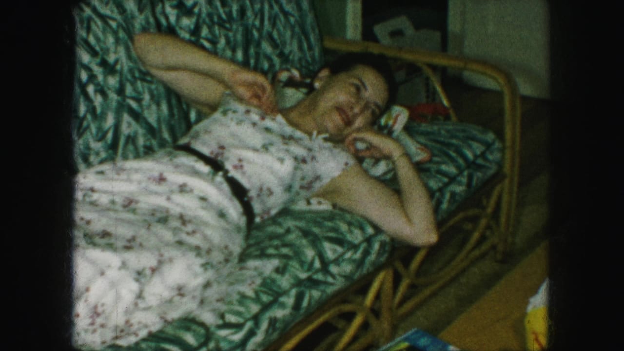 In a 1959 Kansas City home movie, an adult woman reclines on a green patterned couch in a private interior.