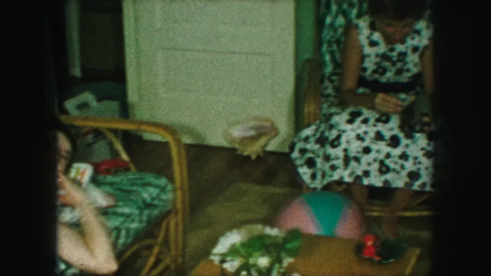 A second adult woman sits beside the couch in a polka-dot dress, handling a small camera in the private room.