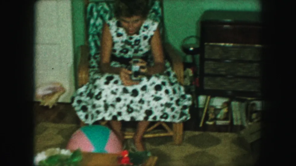 The seated woman looks down at a small camera, with a dresser or cabinet and household objects behind her.