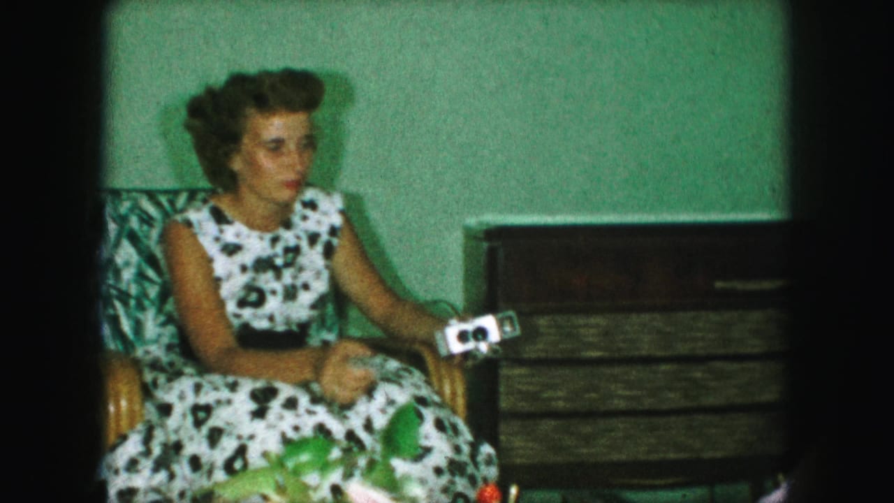 In a 1959 Kansas City home interior, a woman in a black-and-white polka dot dress sits in a wicker chair holding a small camera.
