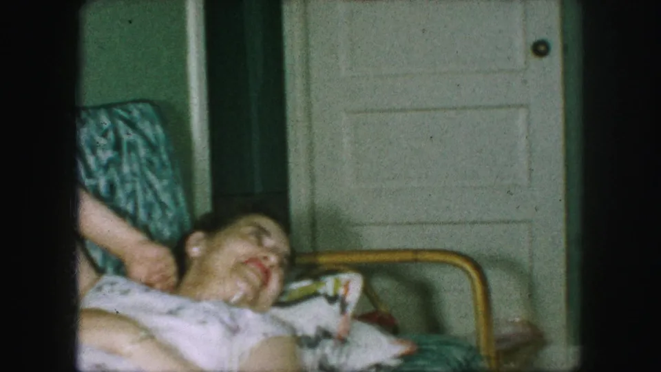 The woman reclines in the living room chair with one arm behind her head, smiling or laughing in the home-movie frame.