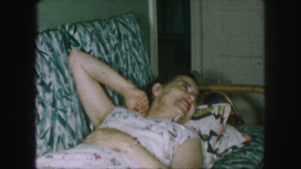 In supported 1959 Kansas City home-movie footage, an adult woman rests on a green-and-white patterned sofa in a floral dress.