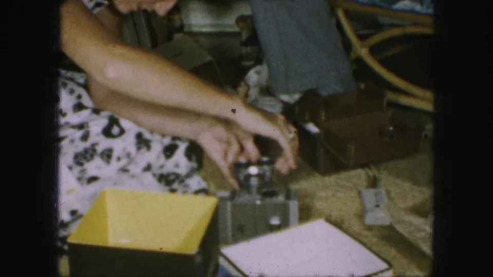 The woman sits or leans forward from the sofa and reaches toward a small vintage camera or device on a nearby table.