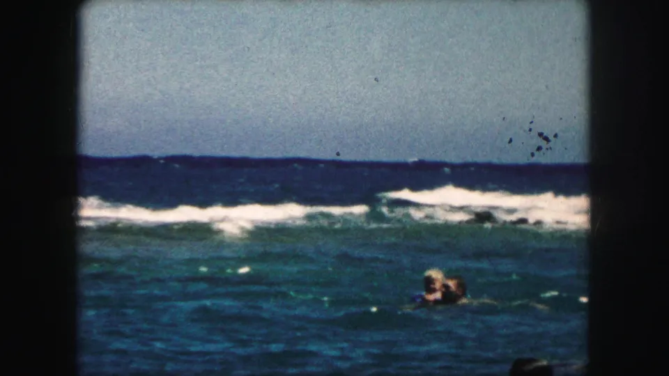 1959 8mm frame of swimmers in ocean water, with low waves, bright sky, and rounded film-frame edges.