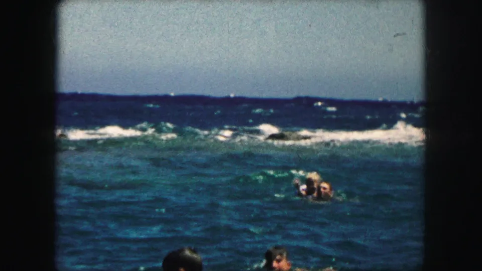 1959 8mm frame of swimmers in blue-green ocean water, with surf bands and a partial foreground swimmer.