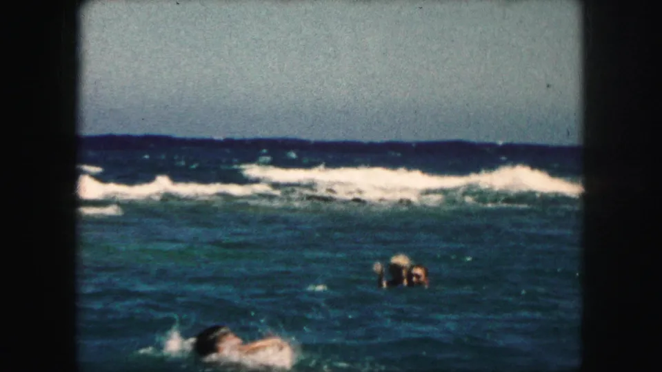 1959 8mm home-movie frame with distant swimmers, white surf, and a foreground swimmer partly crossing the shot.