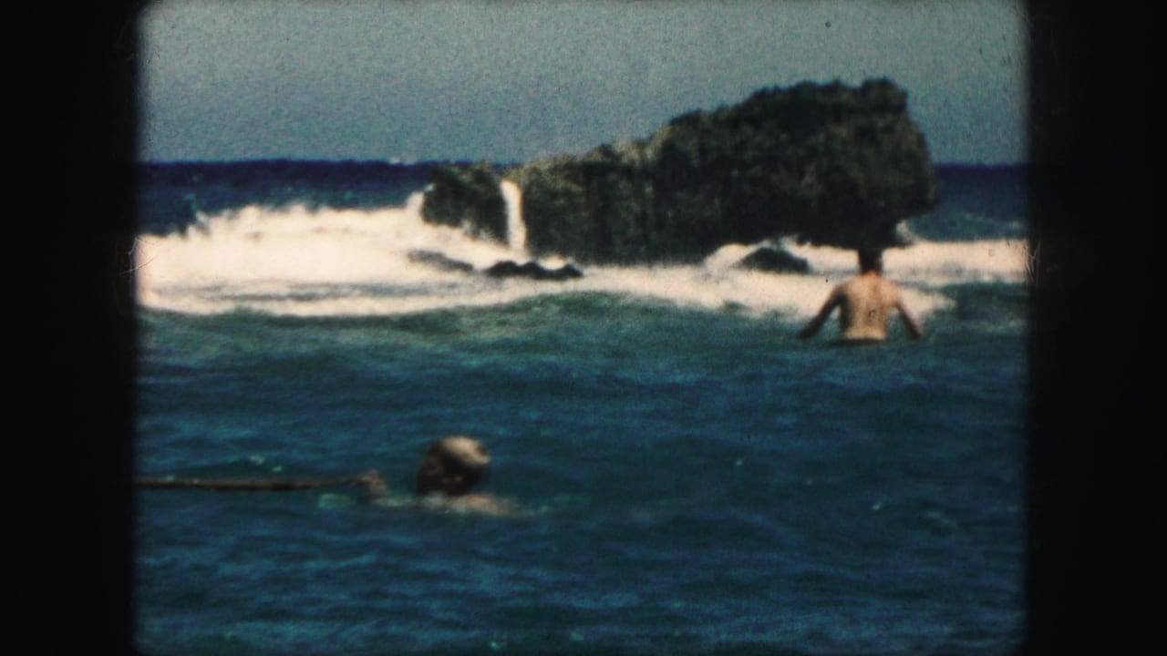Swimmers in blue surf near a dark rocky outcrop with white waves.