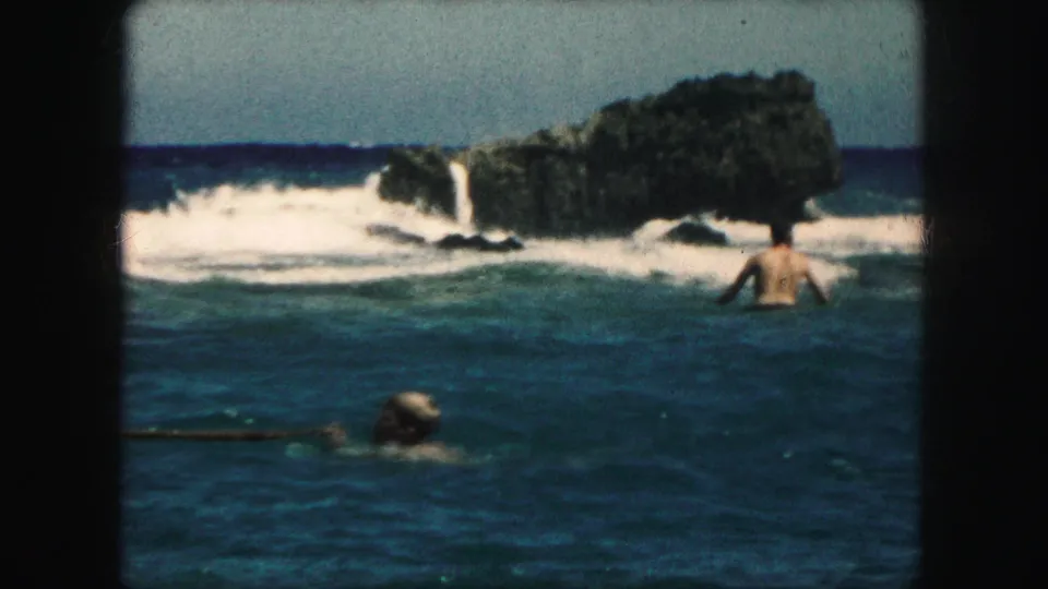 1959 8mm home-movie frame of swimmers in ocean water near a rocky outcrop and breaking surf.